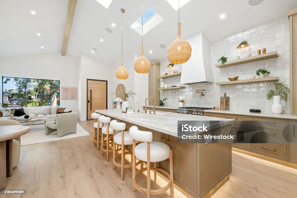 Modern kitchen interior with wooden cabinets, island, and pendant lights, featuring open shelves and a cozy living area.